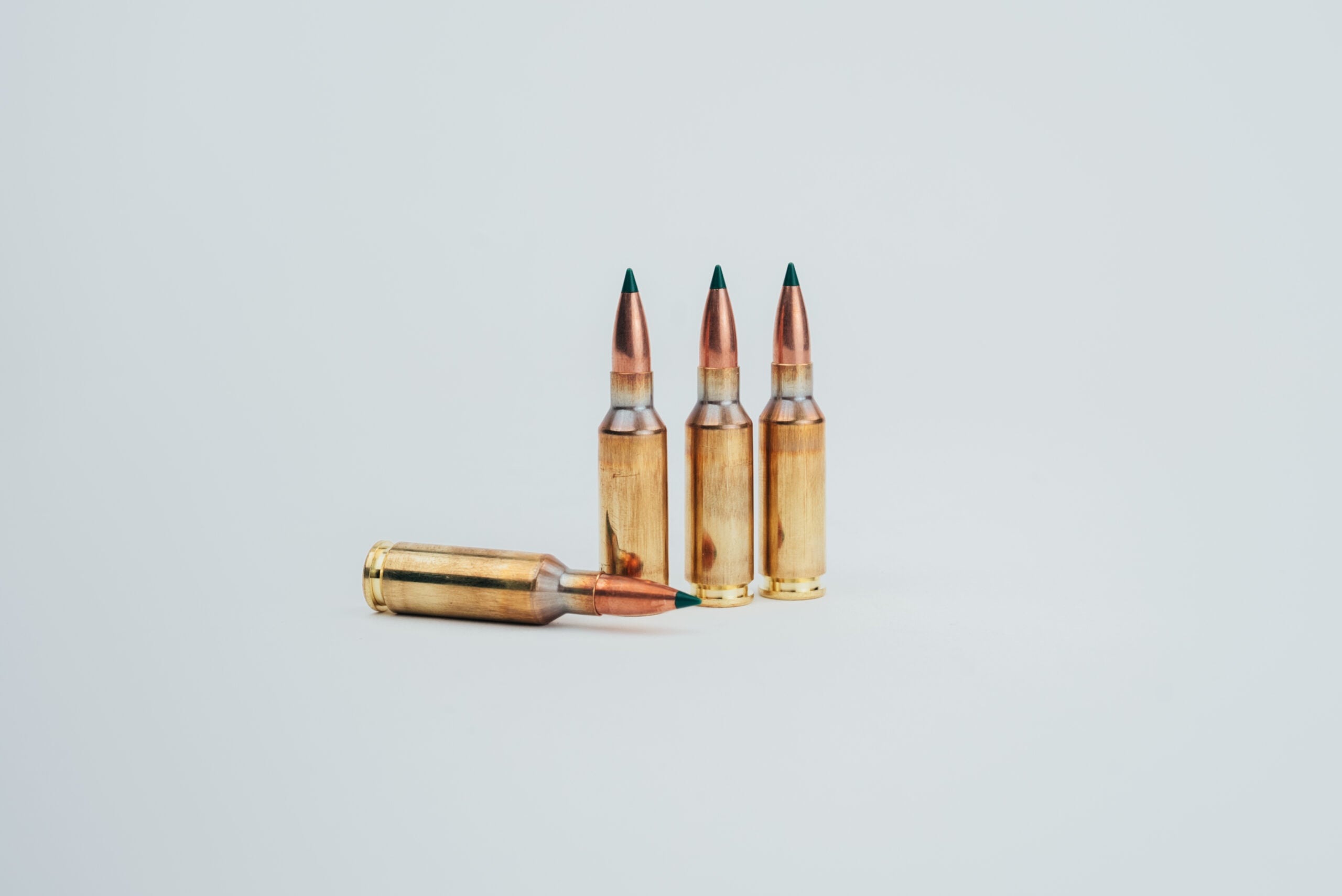 image1 2 - Make Ready Munitions Three copper rifle bullets with green tips standing upright on a light surface, with a fourth cartridge lying to the side.